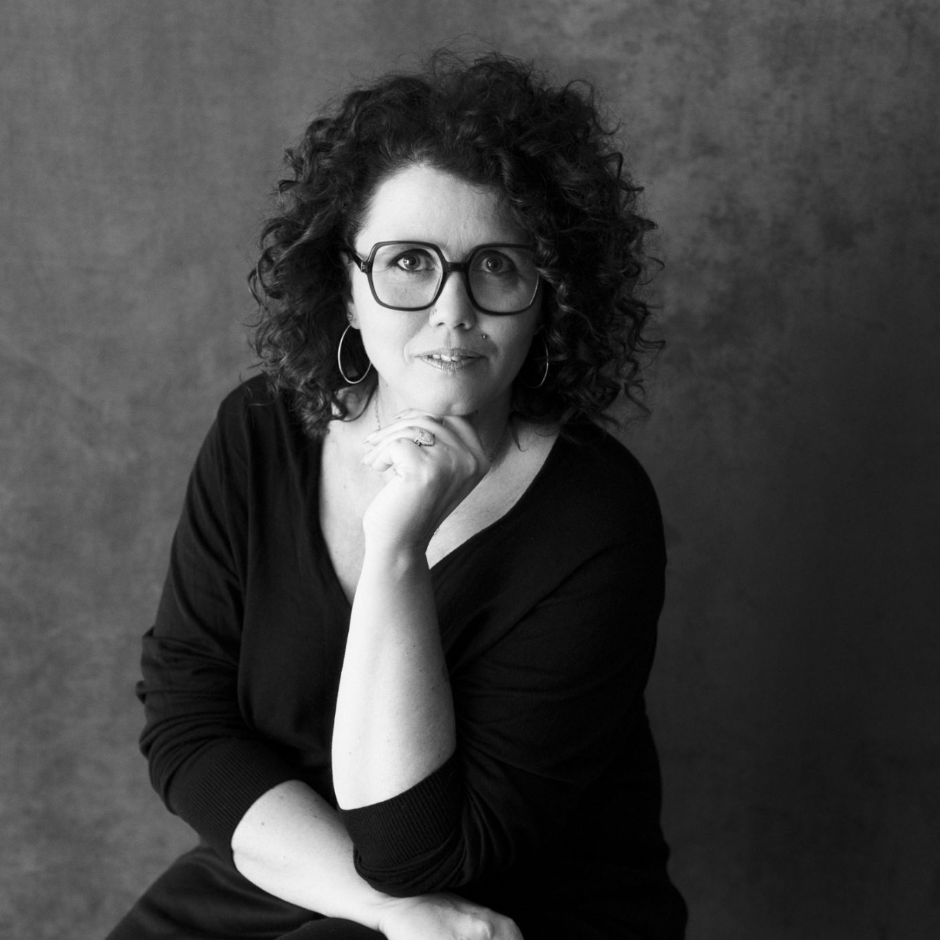 Femme aux cheveux bouclés, portant des lunettes, assise pensivement sur une chaise.